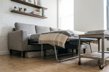Petit salon avec canapé convertible gris déplié révélant son mécanisme de couchage, dans un appartement urbain compact aux murs blancs et parquet chêne clair